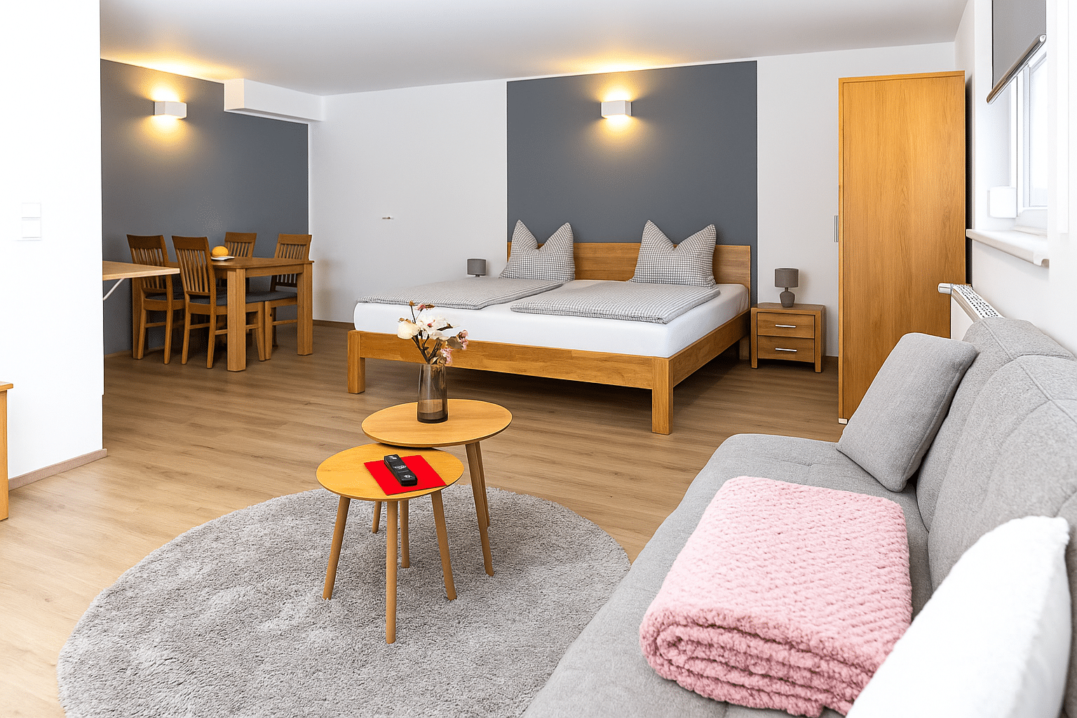 Helles, modernes Studiozimmer mit einem Holzrahmen-Doppelbett vor einer dunkelgrauen Akzentwand, kleinem Nachttisch, Esstisch mit vier Holzstühlen im Hintergrund, grauem Sofa mit rosafarben gefalteter Decke und zwei ineinander stehenden runden Holztischen auf einem runden hellgrauen Teppich, auf denen eine Vase mit Blumen und eine Fernbedienung liegen.