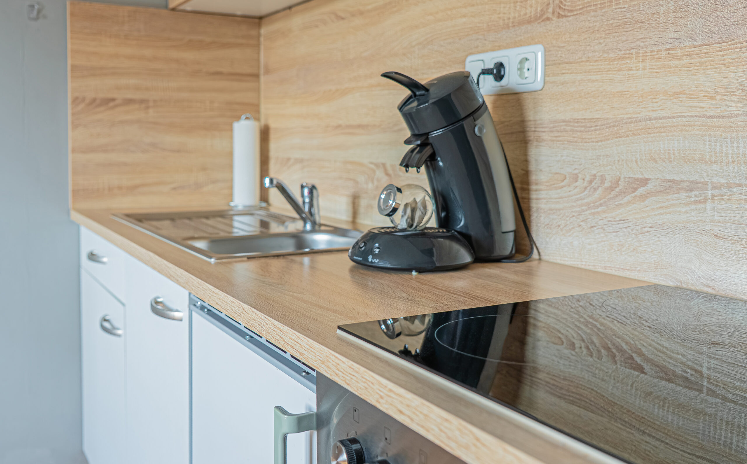 Helle Holzarbeitsplatte in einer Küche mit Edelstahlspüle und Chromarmatur, einer auf der Arbeitsplatte stehenden schwarzen Kaffeemaschine, die an eine Steckdose angeschlossen ist, einem Induktionskochfeld und weißen Unterschränken.