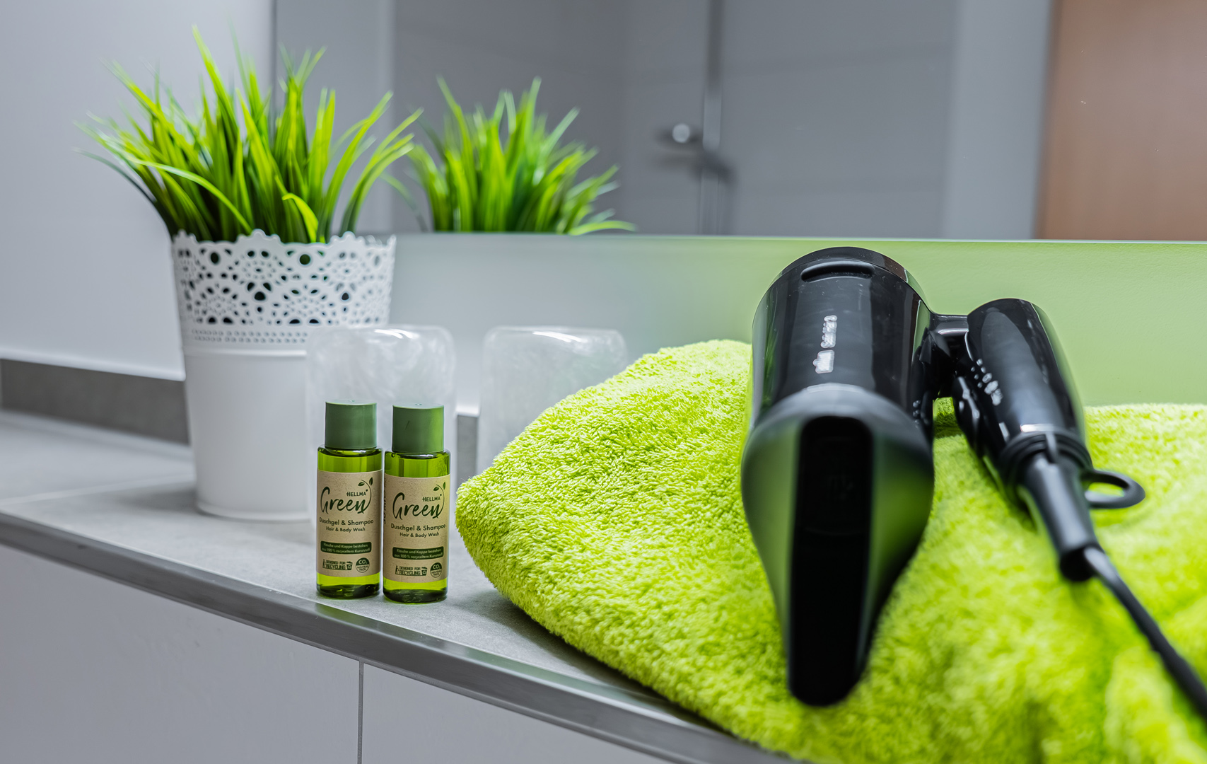Badezimmerablage mit hellgrünem Handtuch, schwarzem Haartrockner, zwei kleinen grünen Shampoo-/Duschgel-Fläschchen und weißem Zierblumentopf vor einem Spiegel.