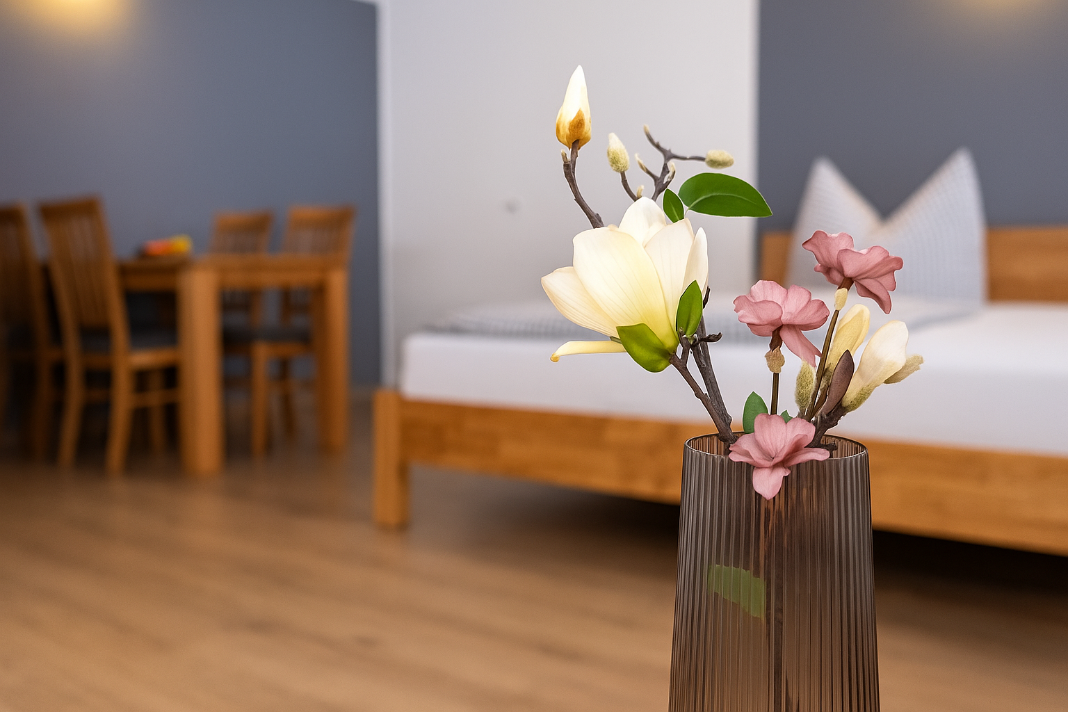 Geriffelte braune Glasvase mit cremefarbenen Magnolien- und zarten rosa Blütenzweigen im Vordergrund, im Hintergrund verschwommenes Holzbett und ein Holz-Esstisch mit Stühlen.