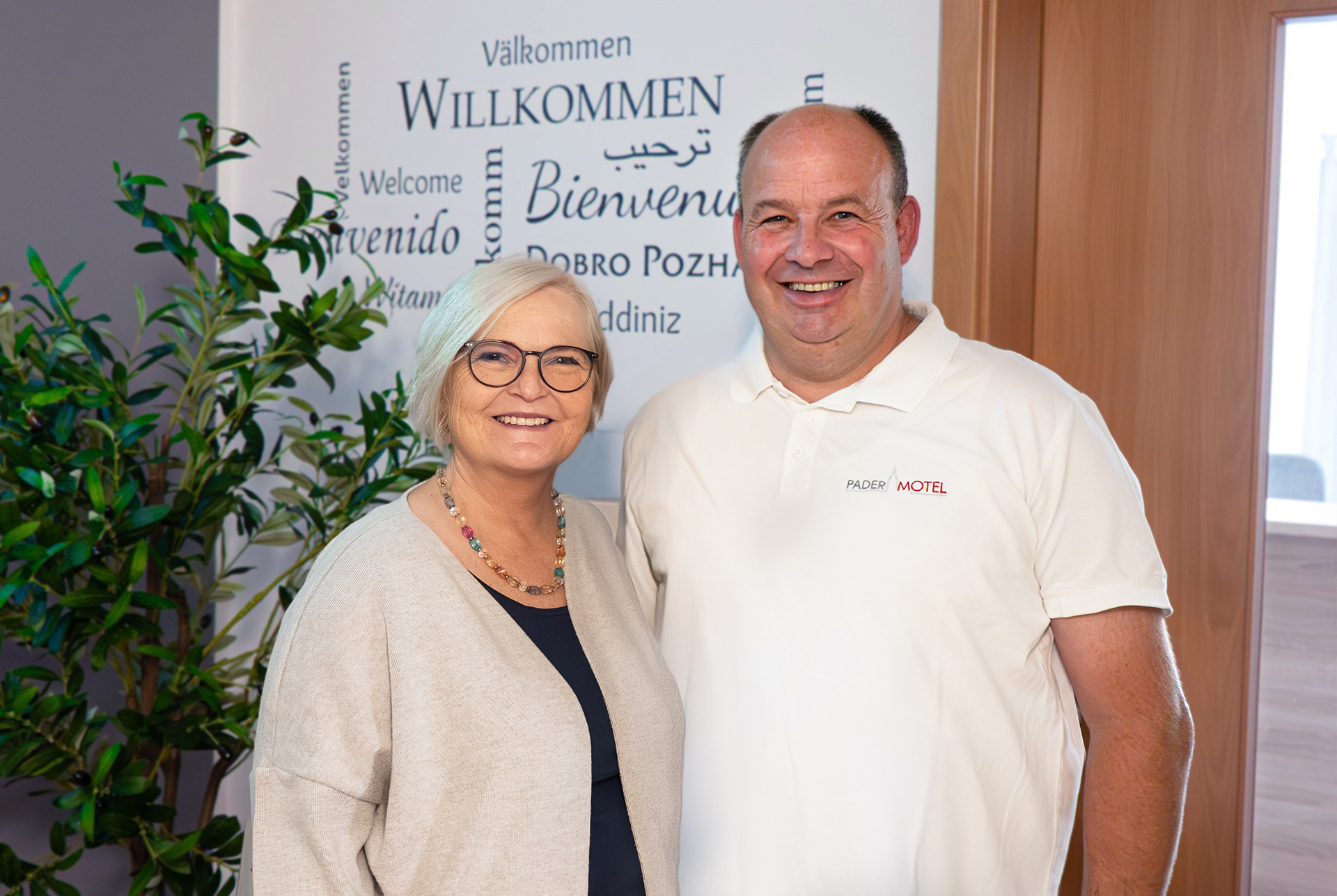 Zwei lächelnde Erwachsene, eine Frau mit kurzen weißen Haaren, Brille und bunter Kette und ein Mann in weißem Polo mit dem Aufdruck 'PADER MOTEL', stehen nebeneinander vor einer Wand mit mehrsprachigem 'Willkommen'-Schriftzug und einer großen Zimmerpflanze.