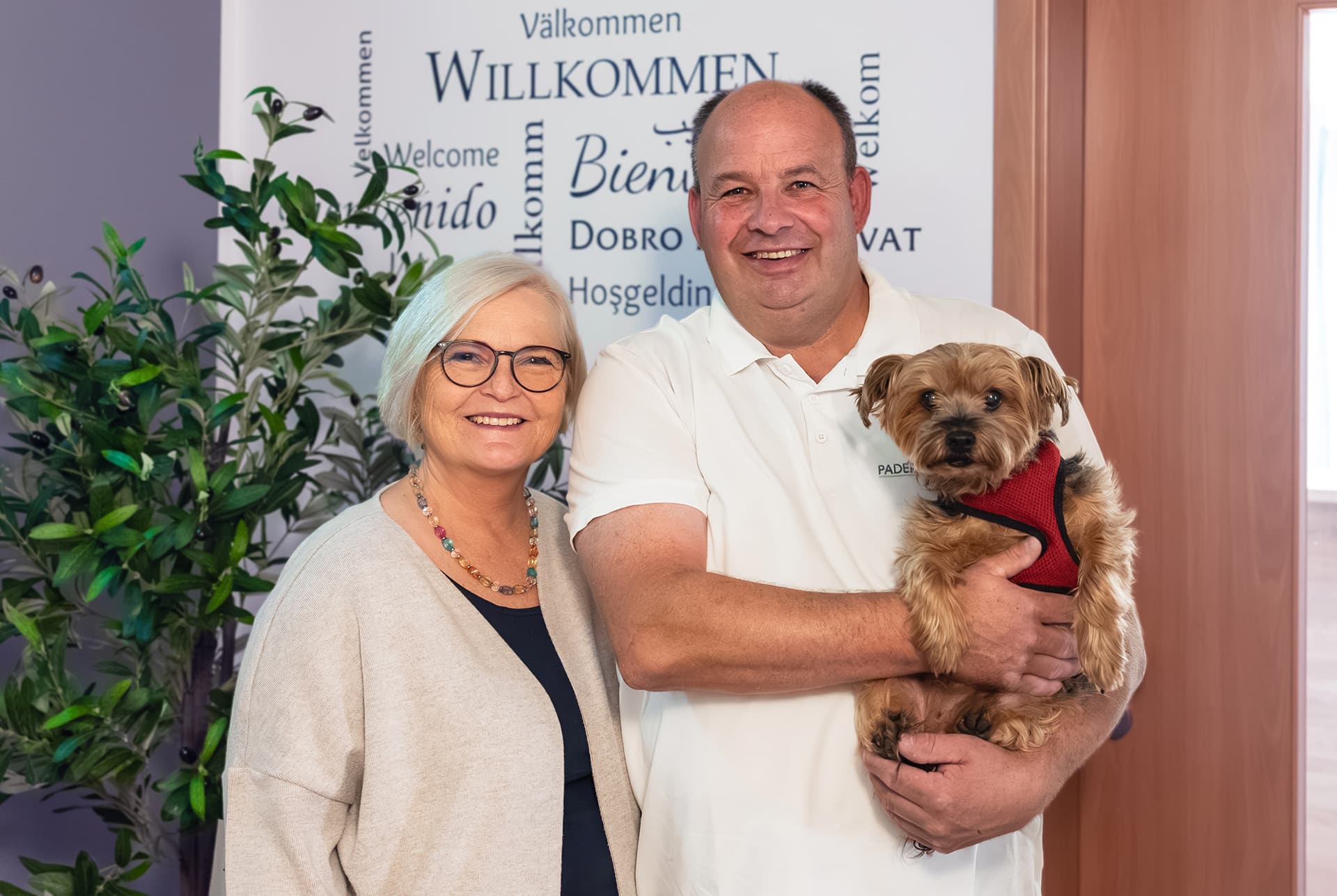 Lächelndes Paar: eine Frau mit kurzweißem Haar und Brille in hellgrauem Cardigan steht neben einem Mann im weißen Polo, der einen kleinen braunen Hund mit rotem Geschirr hält; im Hintergrund eine Wand mit 'Willkommen'-Schrift in mehreren Sprachen und eine Zimmerpflanze.