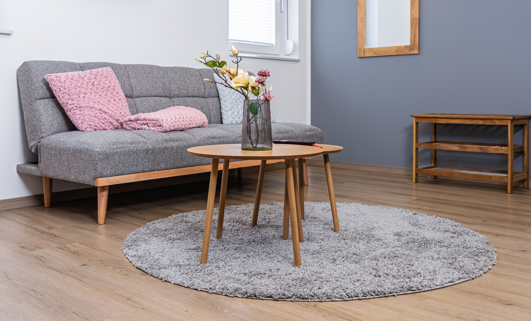 Modernes Wohnzimmer mit grauem Stoffsofa, rosa Strickkissen und -decke, hellem Holz-Couchtisch auf rundem hellgrauen Teppich und geriffelter Glasvase mit gelb-rosa Blumen vor einer blaugrauen Wand und kleiner Holzkonsole.