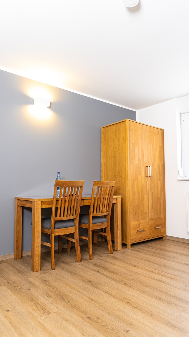 Eckaufnahme eines Zimmers mit hellem Parkettboden, einem massiven Holztisch mit zwei Holzstühlen (graue Sitzpolster) und einer Wasserflasche, daneben ein hoher hölzerner Kleiderschrank vor einer grauen Wand mit heller Wandleuchte.