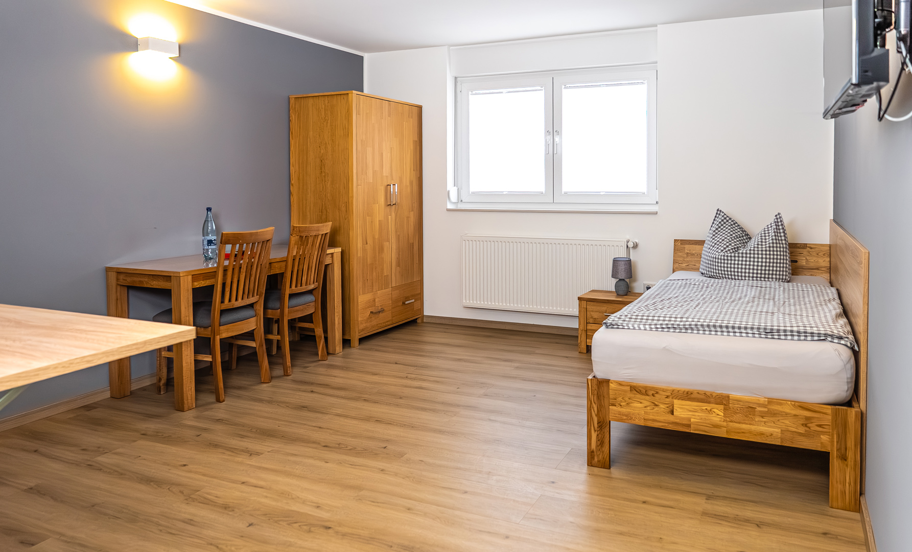 Schlicht eingerichtetes Einzelzimmer mit Holzbett und kariertem Bettzeug rechts, kleinem Nachttisch mit Lampe, Holzkleiderschrank sowie einem Schreibtisch mit zwei Stühlen und Wasserflasche links vor einer grauen Akzentwand und großem Fenster.