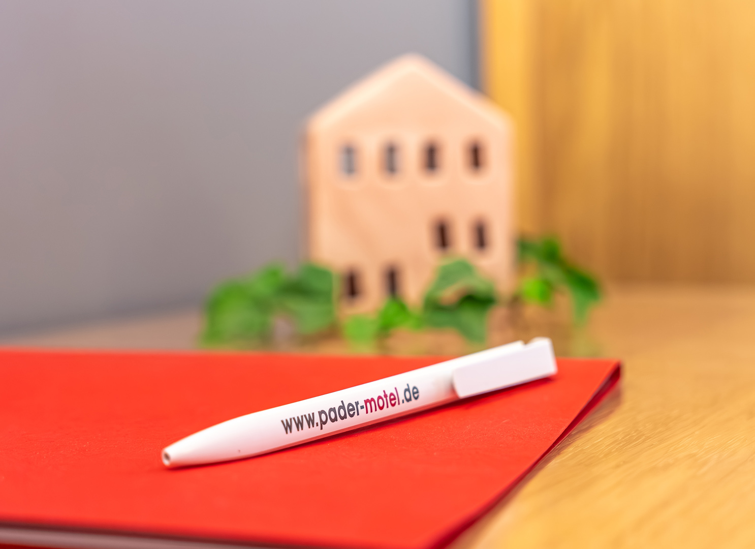 Weißer Kugelschreiber mit der Aufschrift www.pader-motel.de liegt schräg auf einem roten Notizbuch auf einem Holztisch, im unscharfen Hintergrund eine kleine Holzhaus-Dekoration und grüne Pflanze.