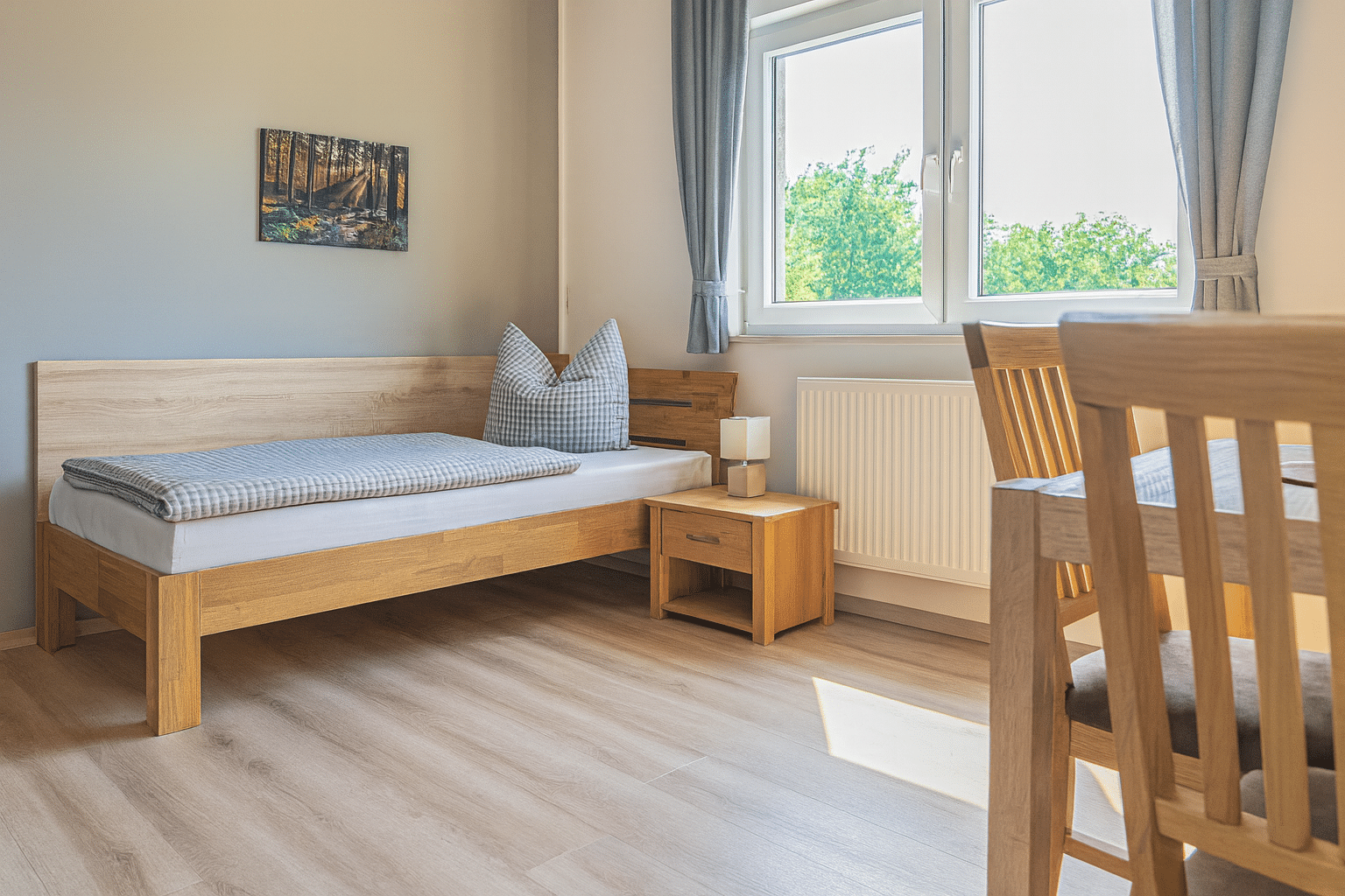 Hell eingerichtetes Schlafzimmer mit einem Einzelbett aus hellem Holz, kariertem grauen Bettzeug und großem Kissen, kleinem Nachttisch mit würfelförmiger Lampe, einem Waldbild an der Wand, großem Fenster mit grauen Vorhängen und Blick auf grüne Bäume sowie hellem Holzboden und teilweiser Ansicht eines Holzstuhls.