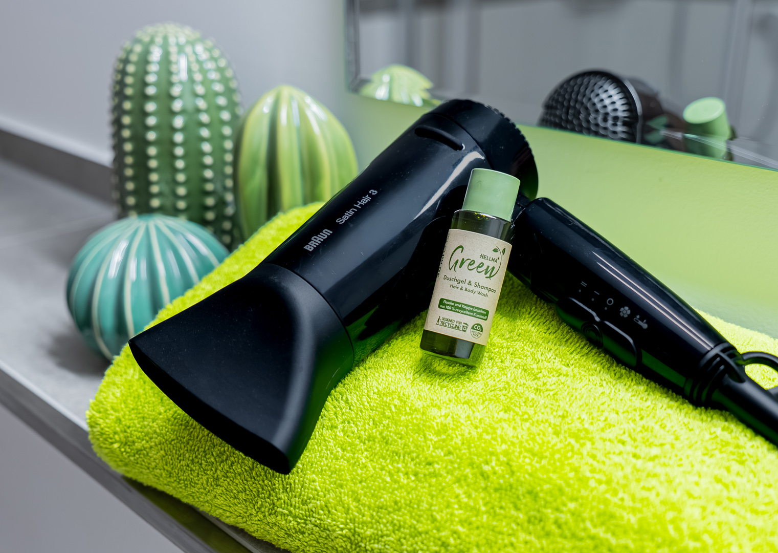 Schwarzer Braun-Haartrockner liegt auf einem leuchtend grünen Handtuch neben einer kleinen Flasche mit der Aufschrift „Green“, im Hintergrund dekorative grüne Keramikkakteen und ein Spiegel.