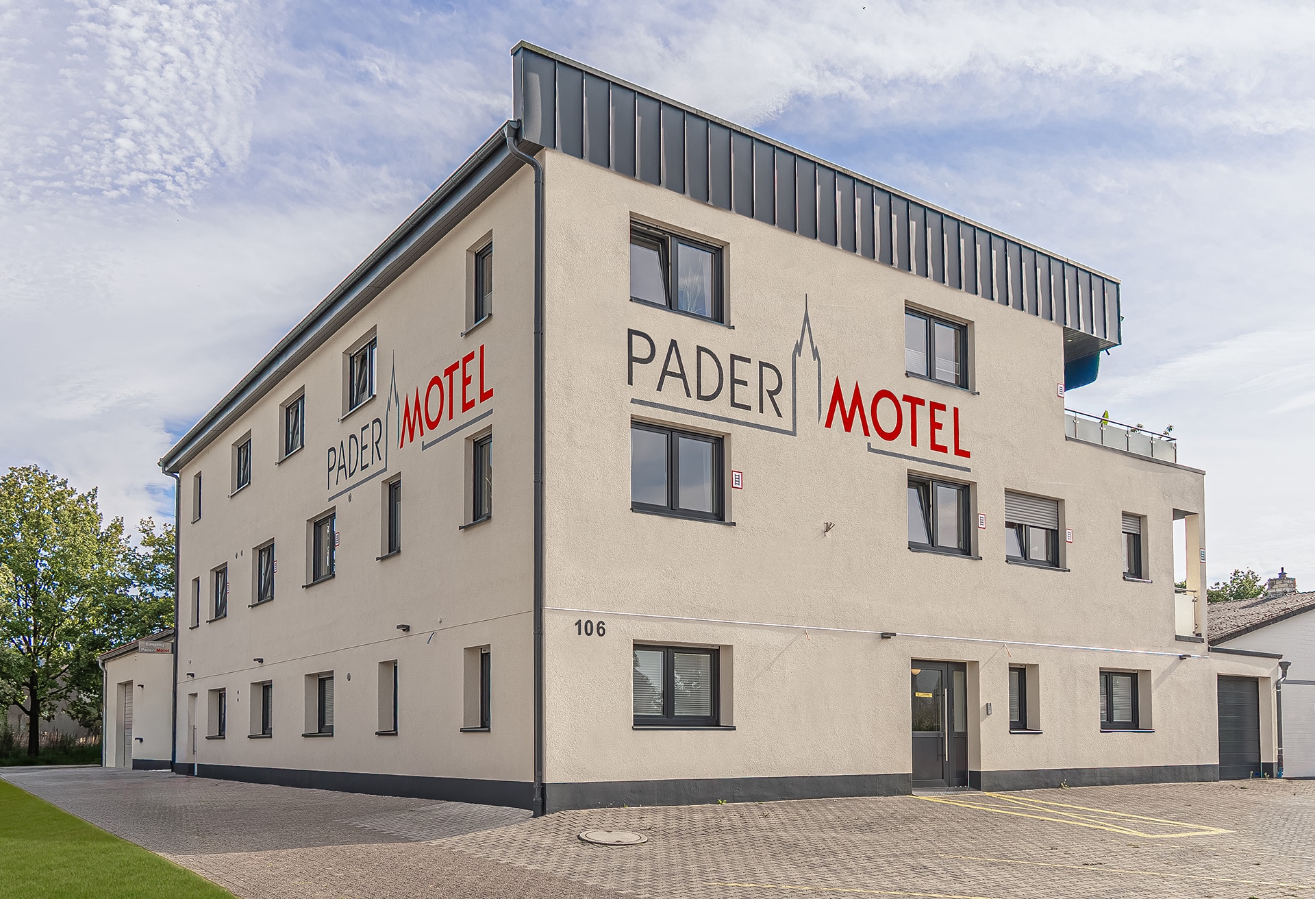Eckaufnahme eines dreistöckigen, beige verputzten Gebäudes mit auffälliger roter und grauer Beschriftung „PADER MOTEL“, mehreren dunklen Fenstern, Parkplatz vor dem Haus und der Hausnummer 106 unter bewölktem Himmel.